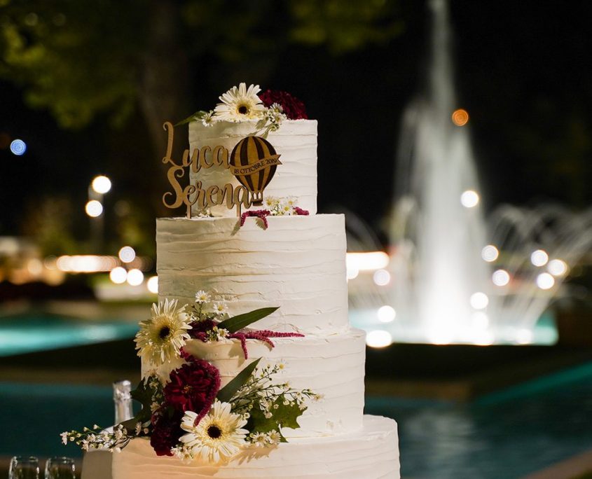 Torta Nuziale Punta Piana Wedding - Twister Adv Torta nuziale a più piani decorata con fiori freschi e topper personalizzato Luca e Serena, con fontana illuminata sullo sfondo per Punta Piana Wedding, realizzata da Twister Adv.