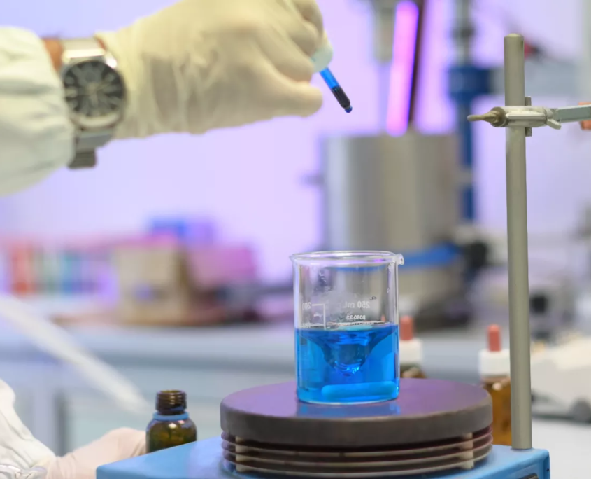 Mano con guanto da laboratorio che versa una soluzione con pipette in un becher con liquido blu su agitatore