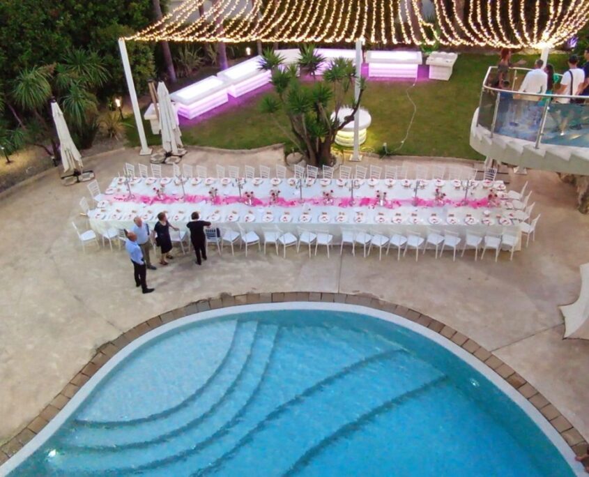 Sala Madame Lilla - Allestimento Ricevimenti e Piscina - Twister ADV Catanzaro Vista dall'alto di una raffinata tavolata imperiale all'aperto allestita a bordo piscina con illuminazione di lucine e banconi luminosi