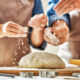 Mani di fornai artigiani che spolverano delicatamente la farina su un panetto di impasto fresco lavorato a mano su un tagliere in legno per Antico Forno Iuliano.