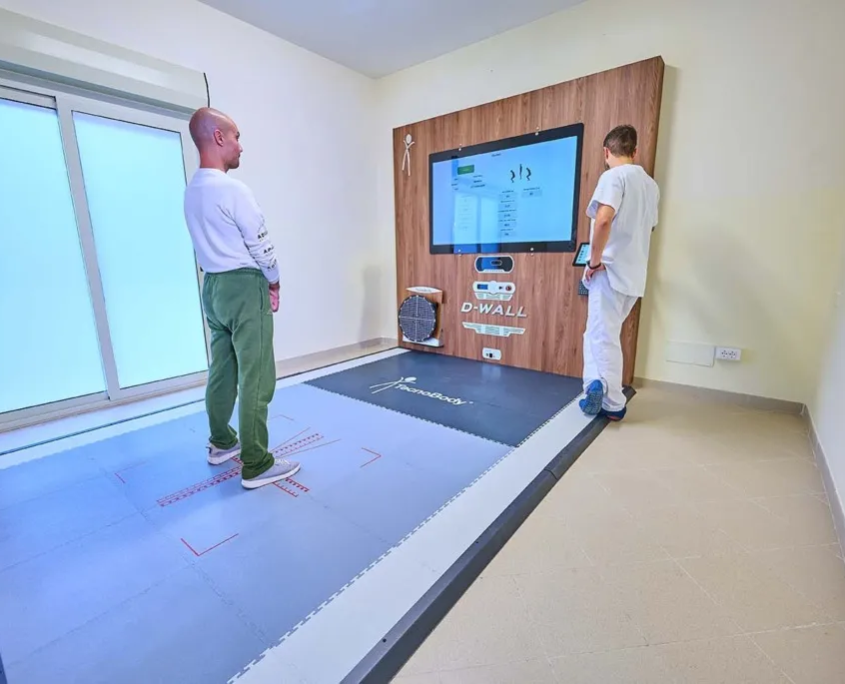 Analisi Posturale e Valutazione Cinematica D-Wall TecnoBody - Parisan Catanzaro Paziente posizionato su pedana stabilometrica di fronte allo specchio digitale interattivo D-Wall TecnoBody per analisi posturale, assistito da un fisioterapista nel centro Parisan.